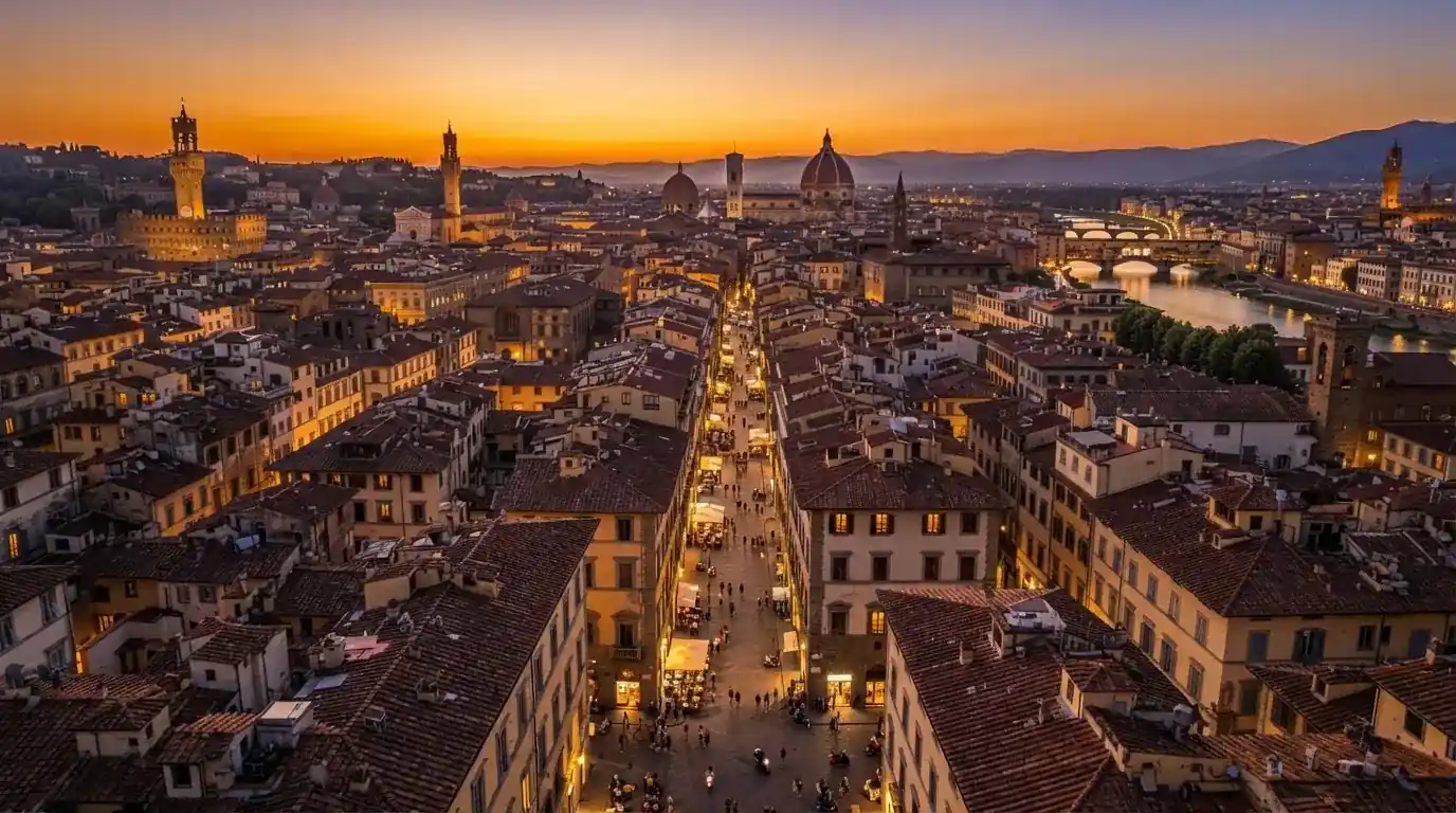 Vista dall'alto di una città italiana al tramonto, con luci che si accendono nelle finestre degli edifici lungo una via trafficata.